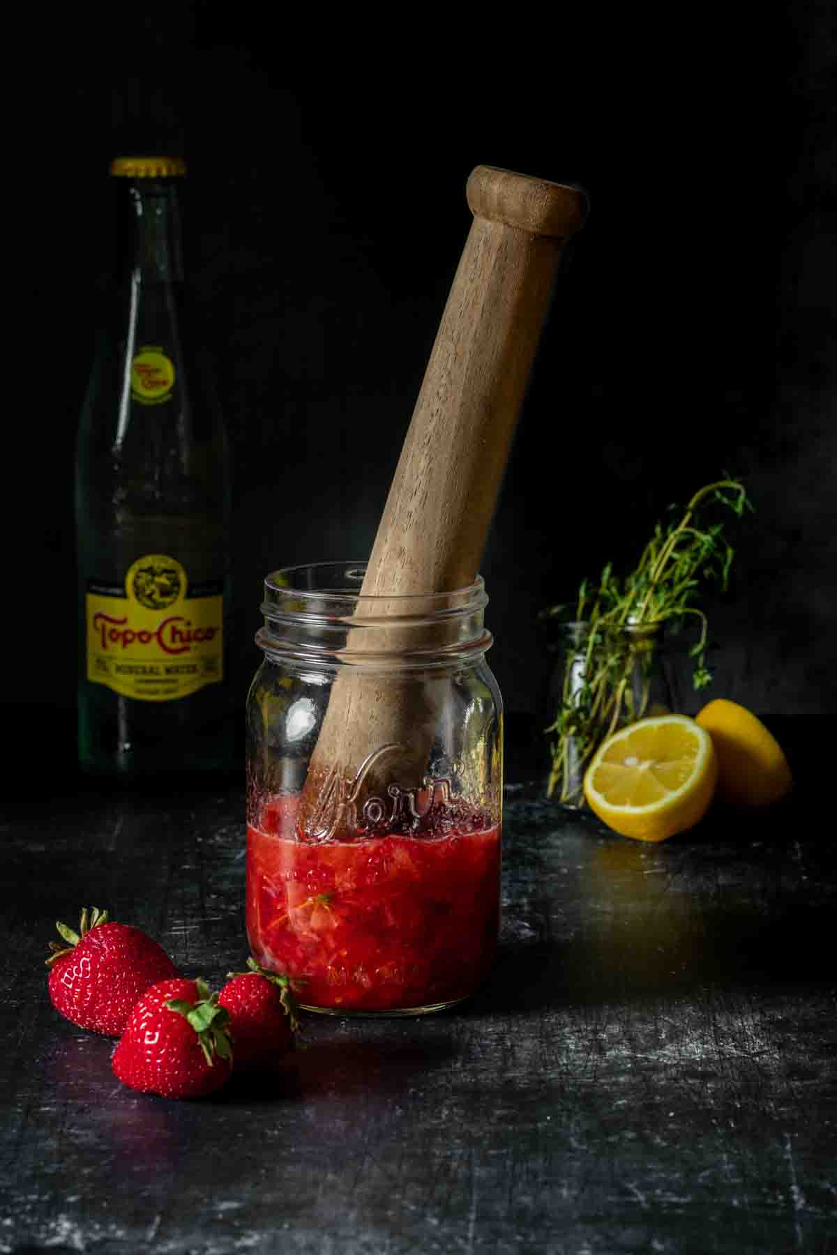 A muddler mashing strawberries, thyme, lemon juice, vodka and sugar in a jar