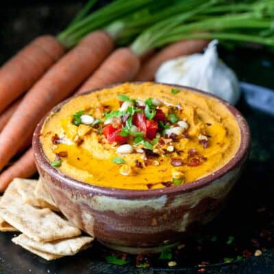 A brown bowl filled with roasted carrot hummus on a dark surface