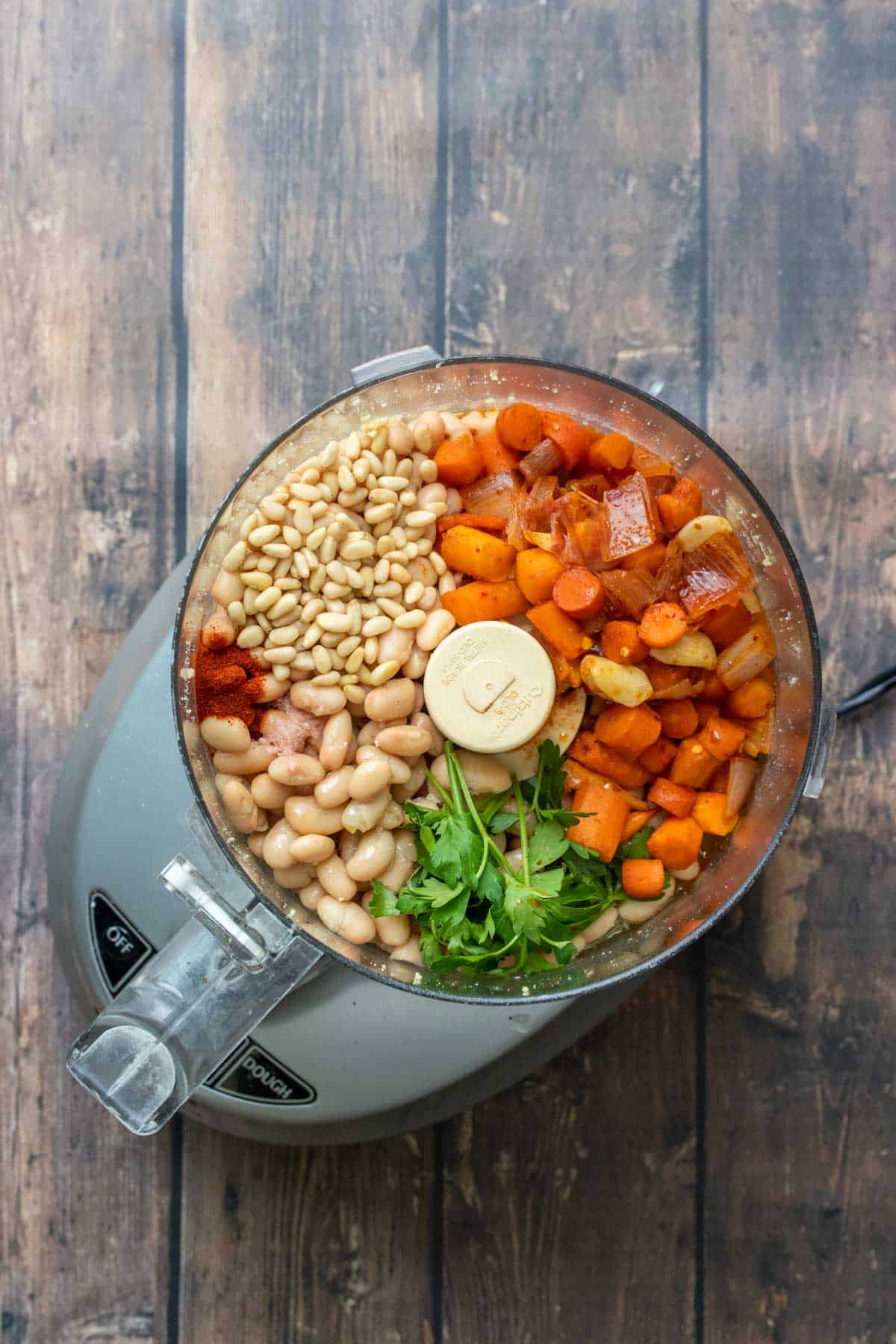 Top view of a food processor with pine nuts, beans, carrots and parsley inside