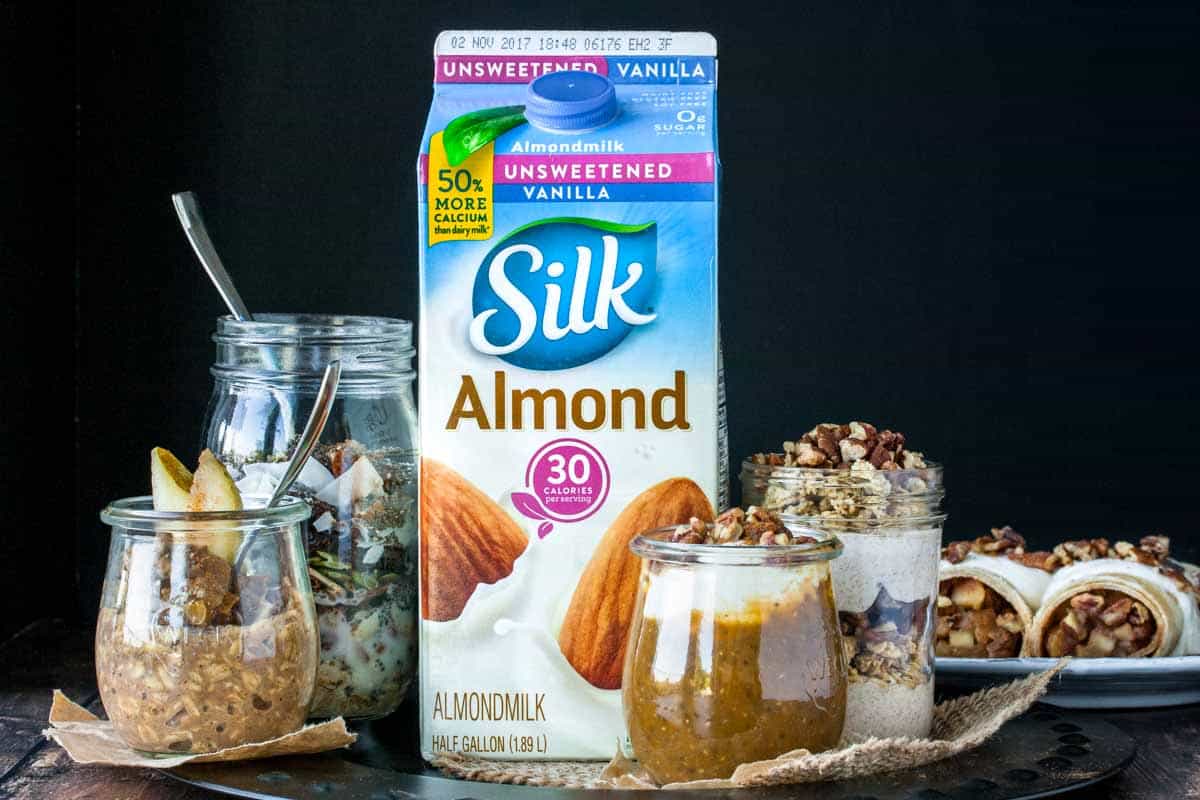 A colorful carton of Silk Almond milk surrounded by a collection of breakfast type foods like oats, chia pudding, yogurt and crepes.