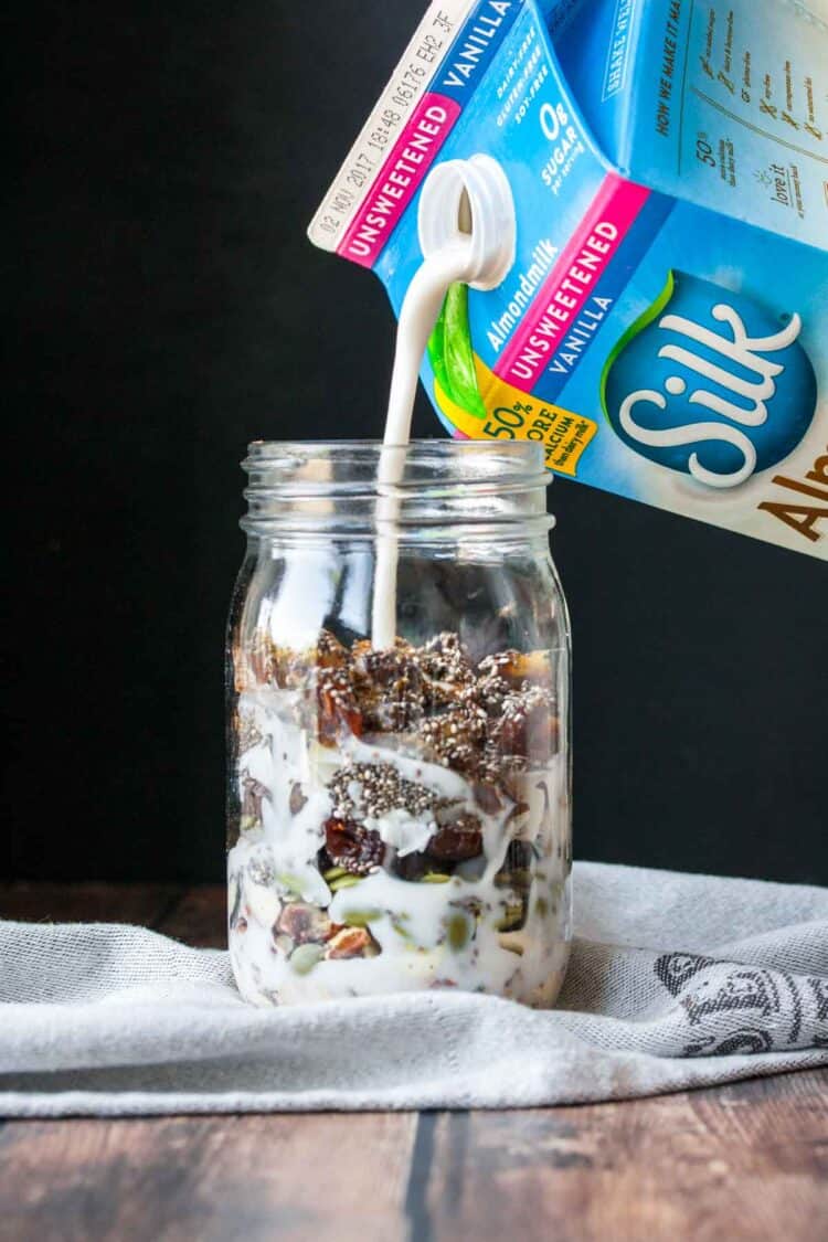 A blue and pink carton of Silk almond milk pouring milk into a glass jar with homemade cereal inside.