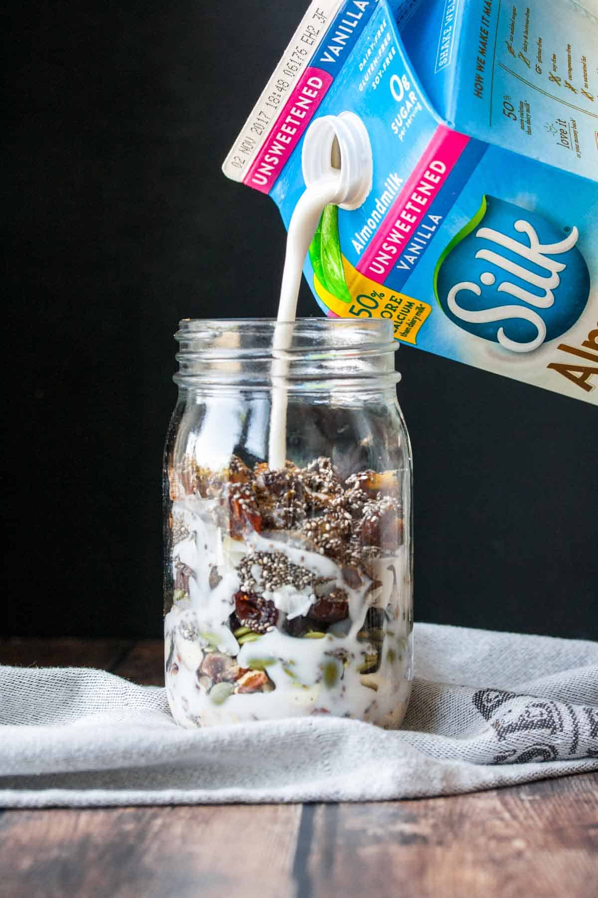 A blue and pink carton of Silk almond milk pouring milk into a glass jar with homemade cereal inside.