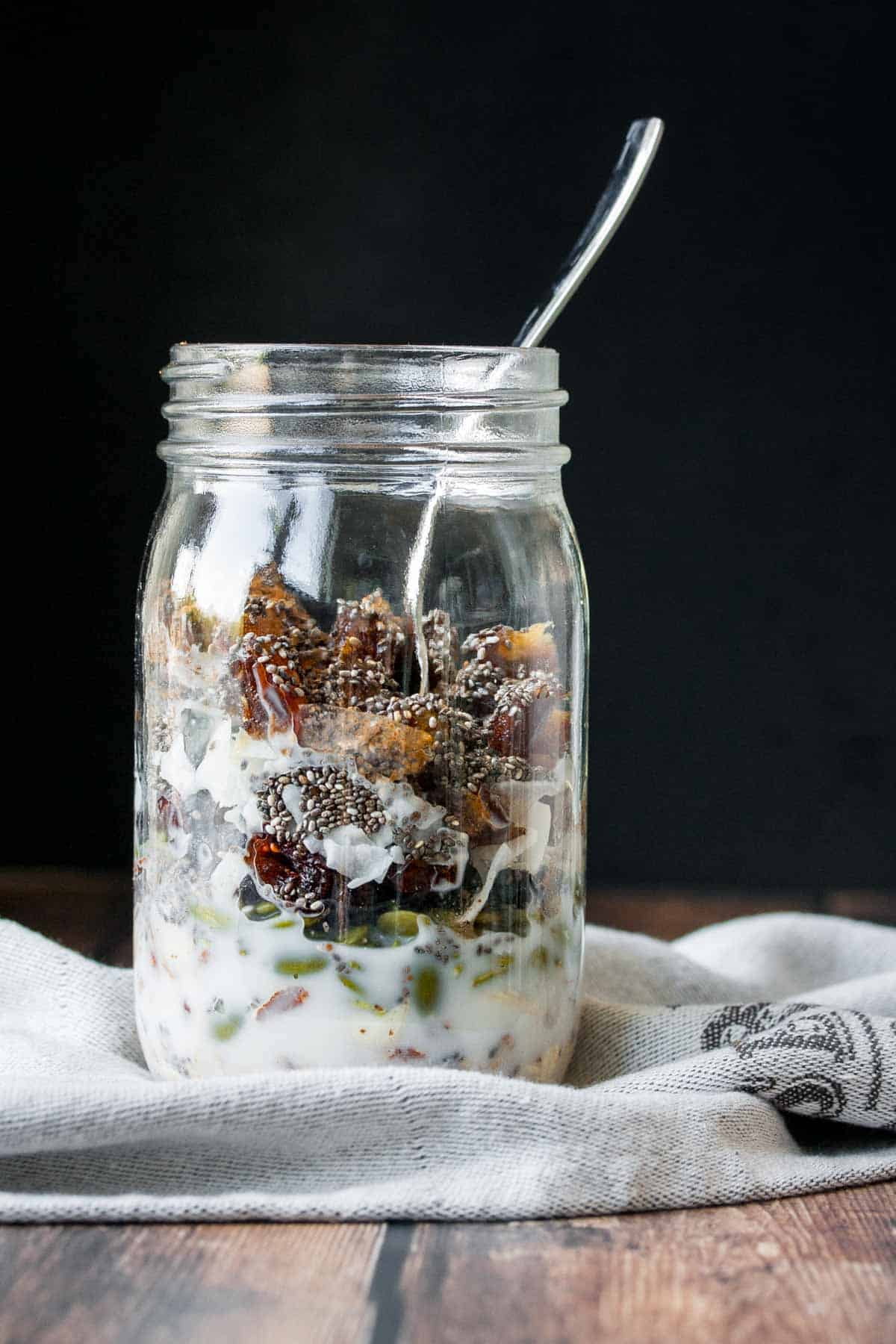 A glass jar with a homemade cereal of dried fruits, nuts and oats and with a little milk on the bottom and a spoon in it.
