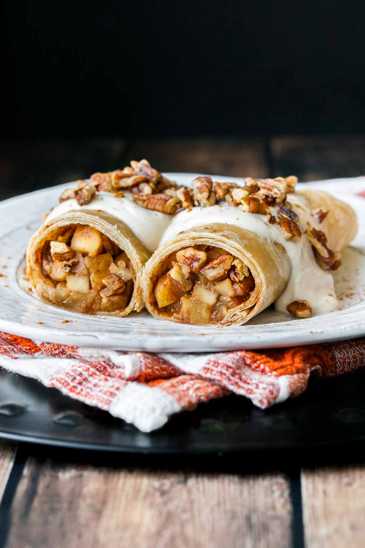 A white plate on an orange checkered towel with apple filled rolled crepes and topped with nuts.