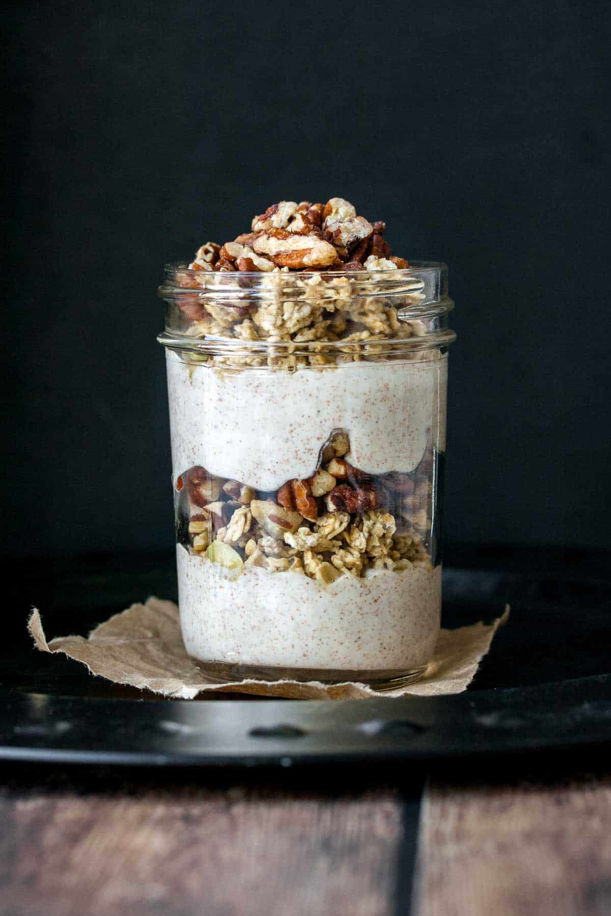 A glass jar layered with yogurt and granola and topped with nuts sitting on a piece of brown paper.