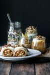 Chia pudding, oats, yogurt parfait and homemade cereal in jars behind a white plate with apple filled crepes on a wooden table.