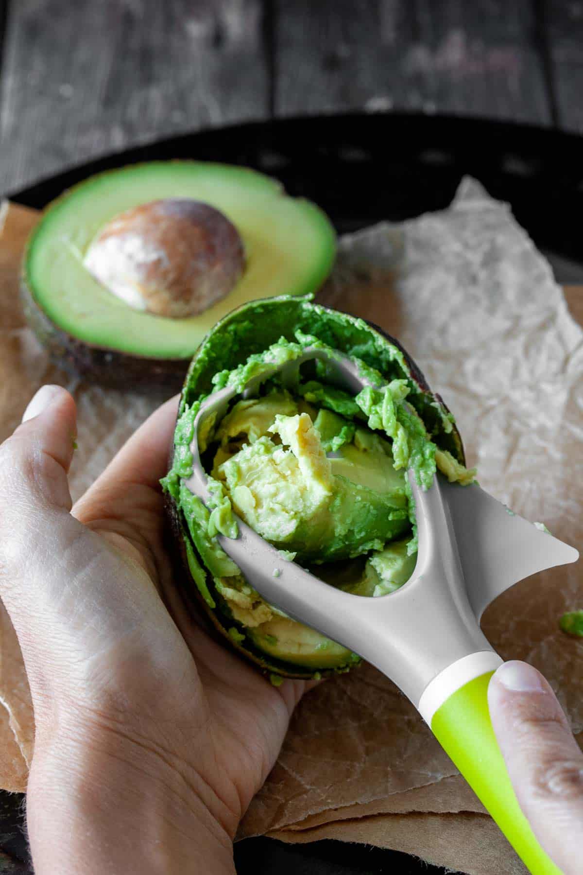 Kitchen tool scraping avocado from a half avocado being held in the palm of a hand.
