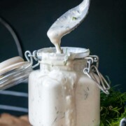 A spoon dripping white tzatziki sauce into a glass jar full of it sitting on parchment