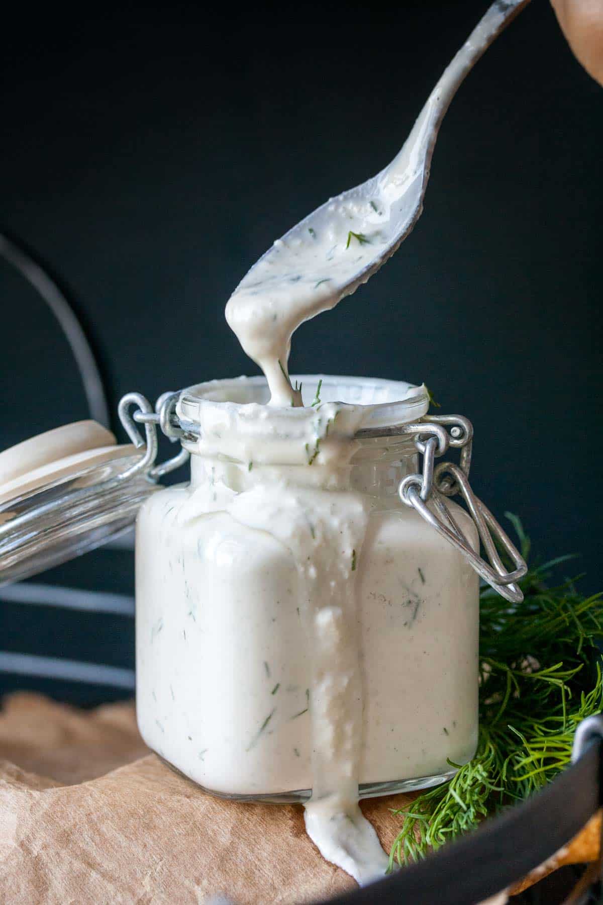 A spoon dripping white tzatziki sauce into a glass jar full of it sitting on parchment