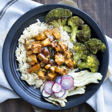 Black plate with General Tso's tofu, roasted broccoli, fennel, and roasted radishes on top of brown rice