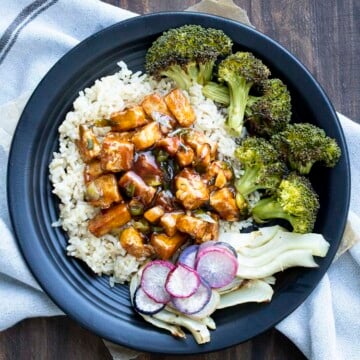 Black plate with General Tso's tofu, roasted broccoli, fennel, and roasted radishes on top of brown rice