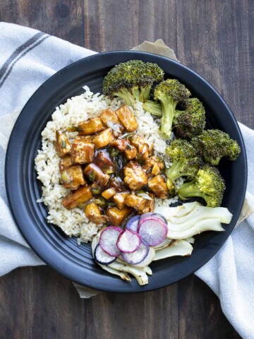 Black plate with General Tso's tofu, roasted broccoli, fennel, and roasted radishes on top of brown rice