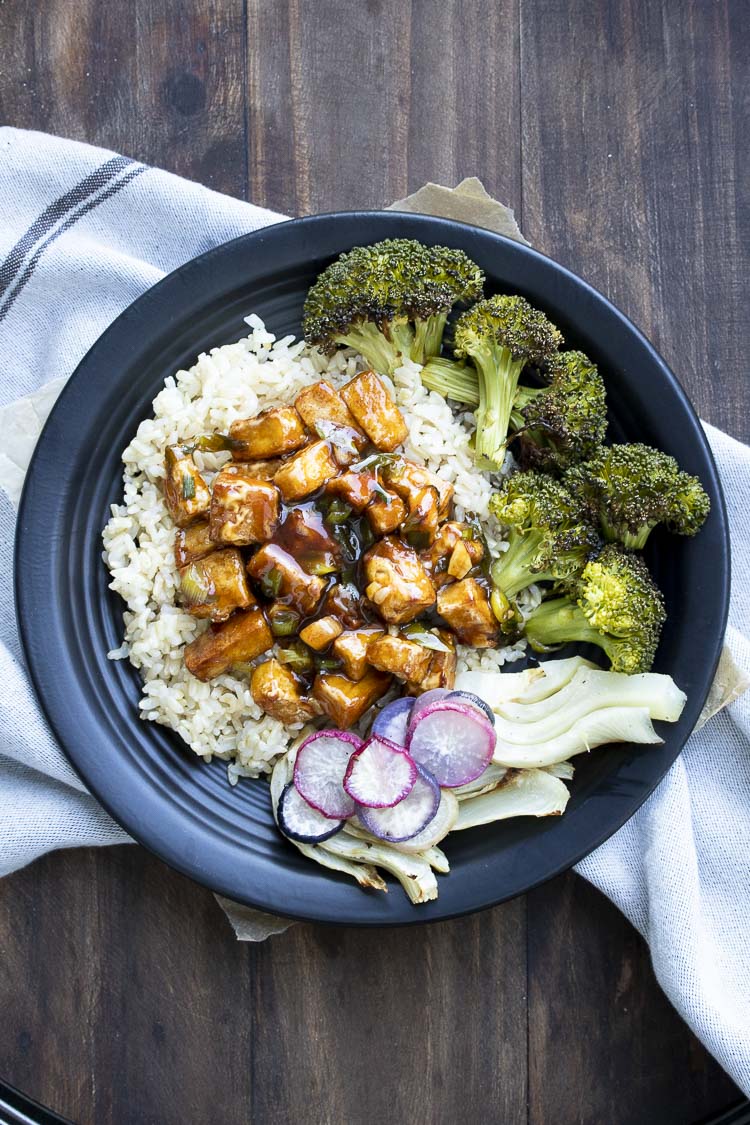 Black plate with General Tso's tofu, roasted broccoli, fennel, and roasted radishes on top of brown rice