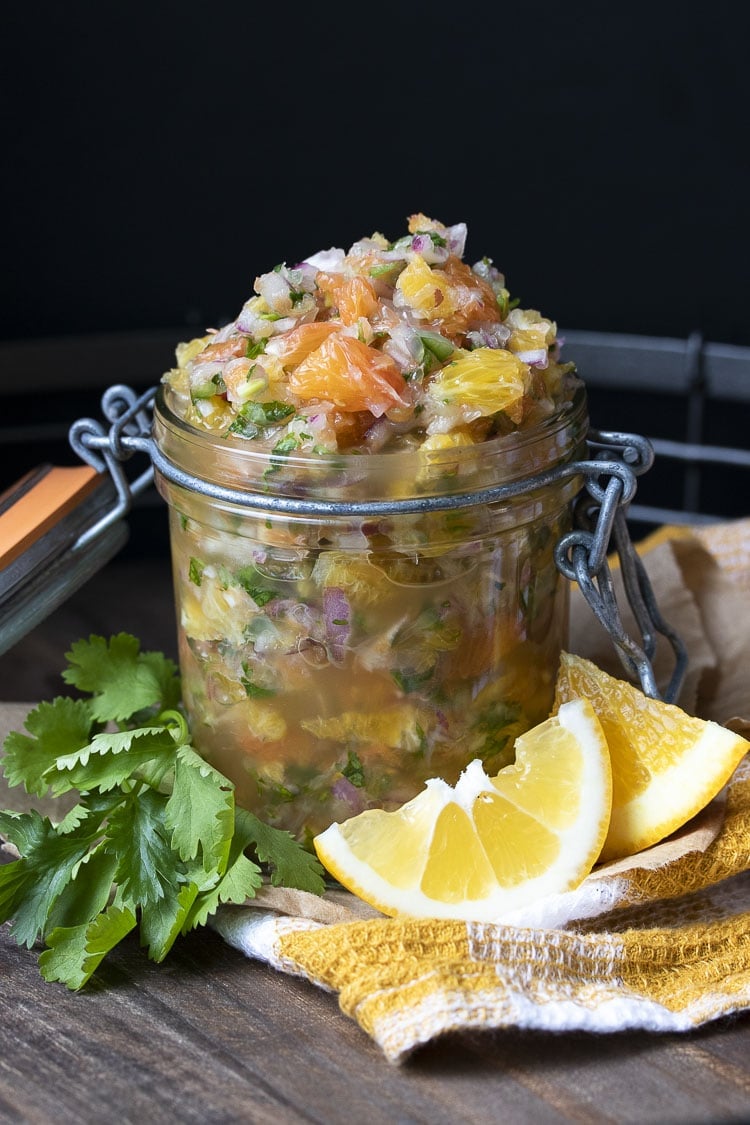 Glass jar of fresh citrus salsa on a wooden tray next to cilantro and orange slices