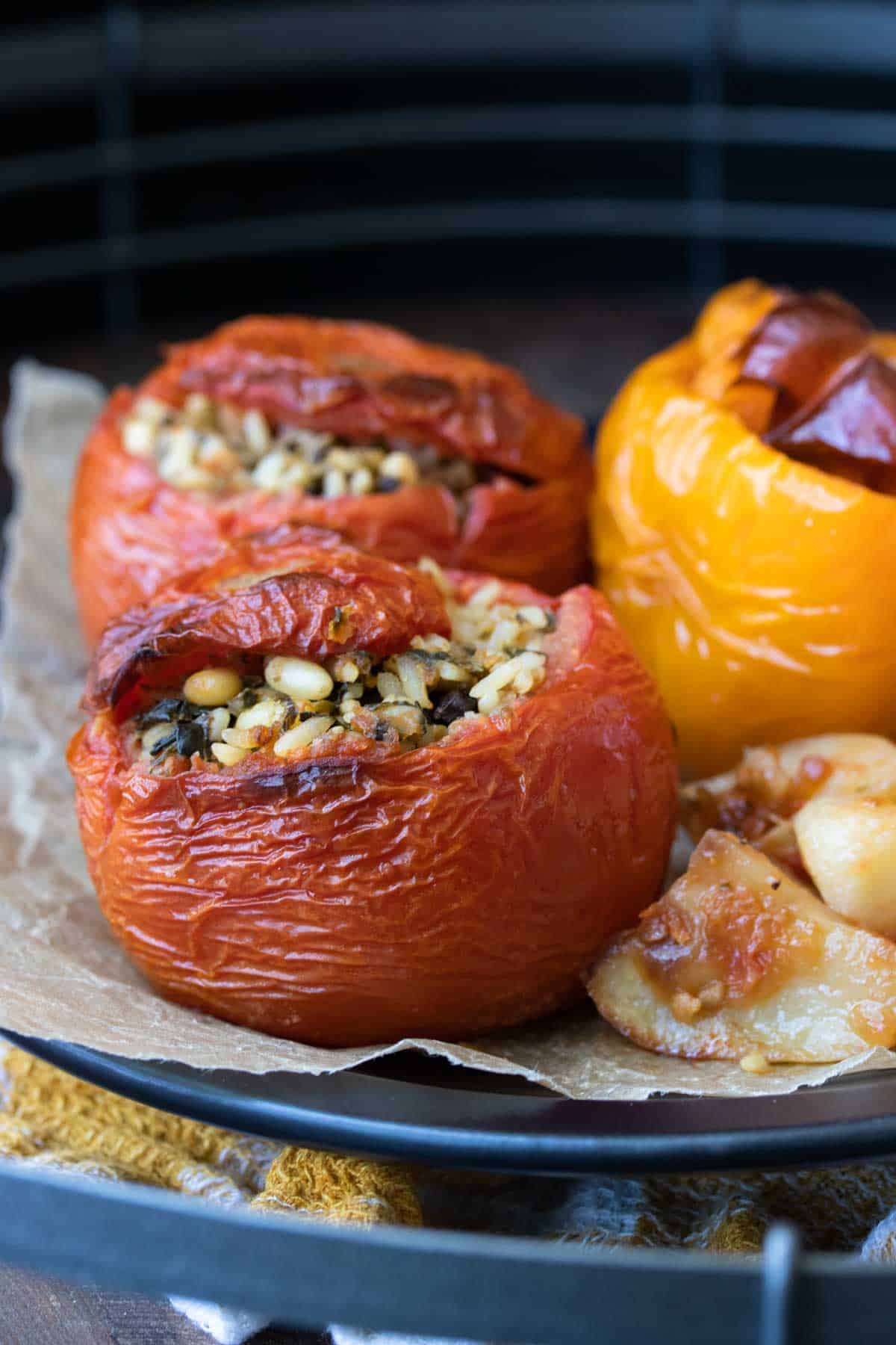 Tomatoes and peppers stuffed with a rice mixture next to pieces of potato on a piece of parchment sitting on a black plate.