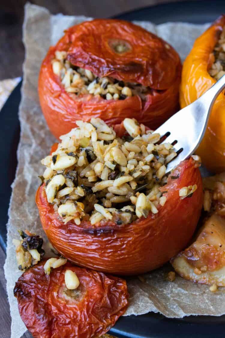 Fork scooping rice from a baked stuffed tomato on a black plate with others.