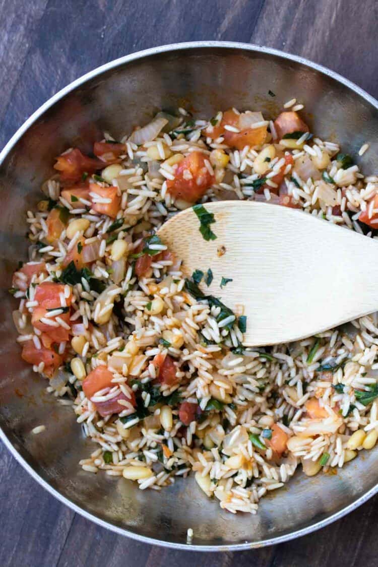 Wooden spoon stirring rice mixture with tomatoes and herbs cooking in a stainless steel pan.