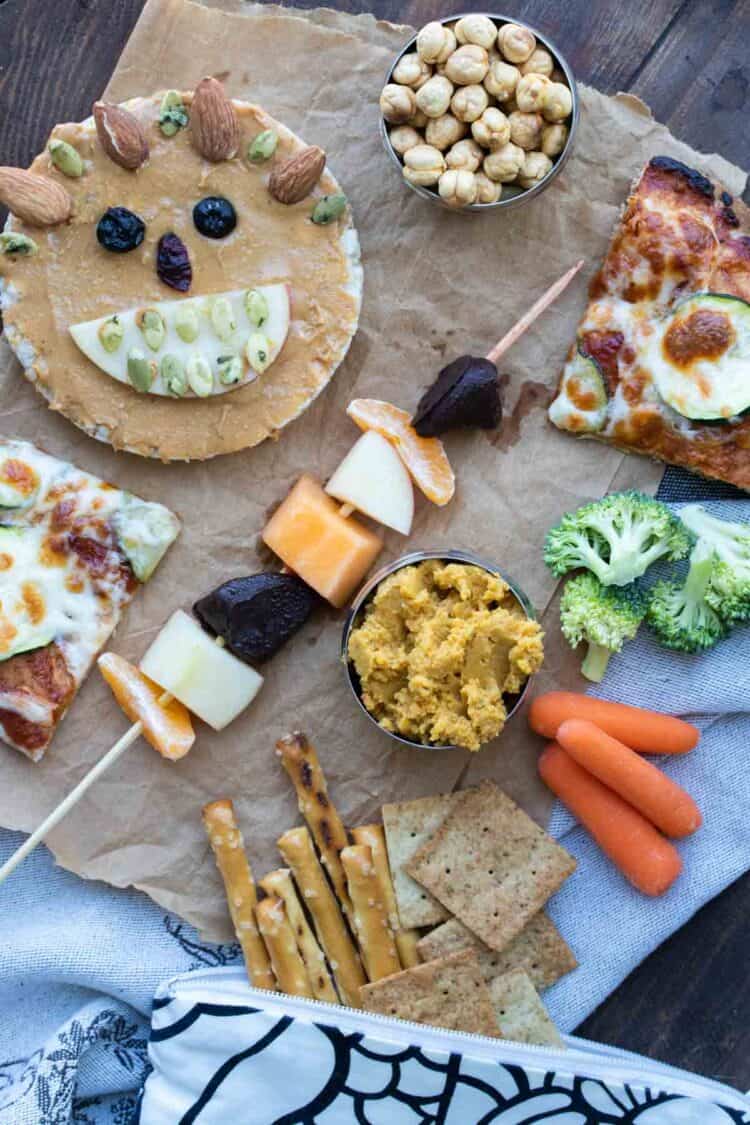 A variety of school lunch items like fruit kebabs, pizza, hummus, veggies and pretzels.