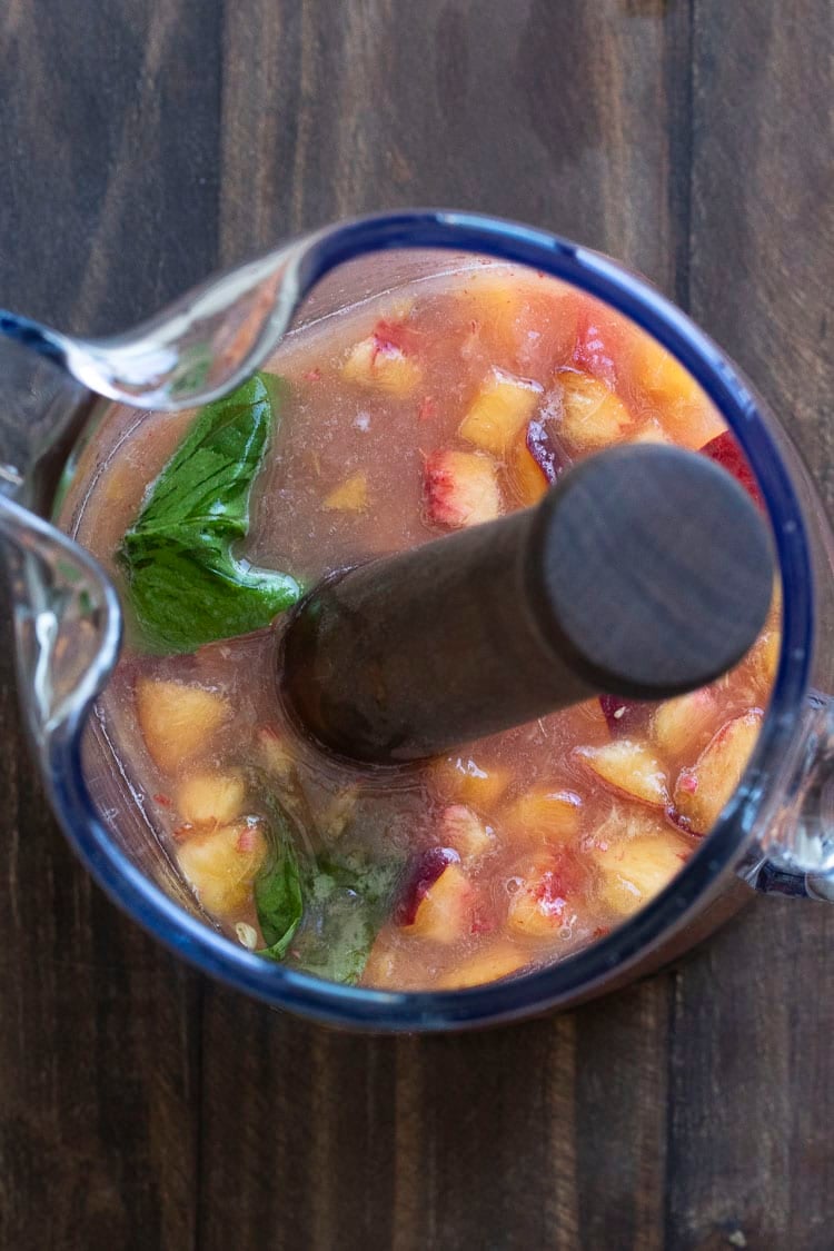 Top view of glass pitcher with grapefruit juice, basil and chopped peaches being mixed