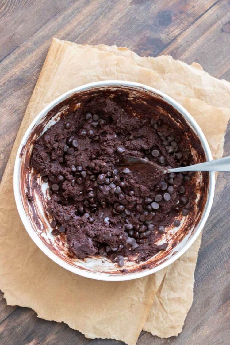 Metal spoon mixing chocolate chips into brownie batter in a while bowl.