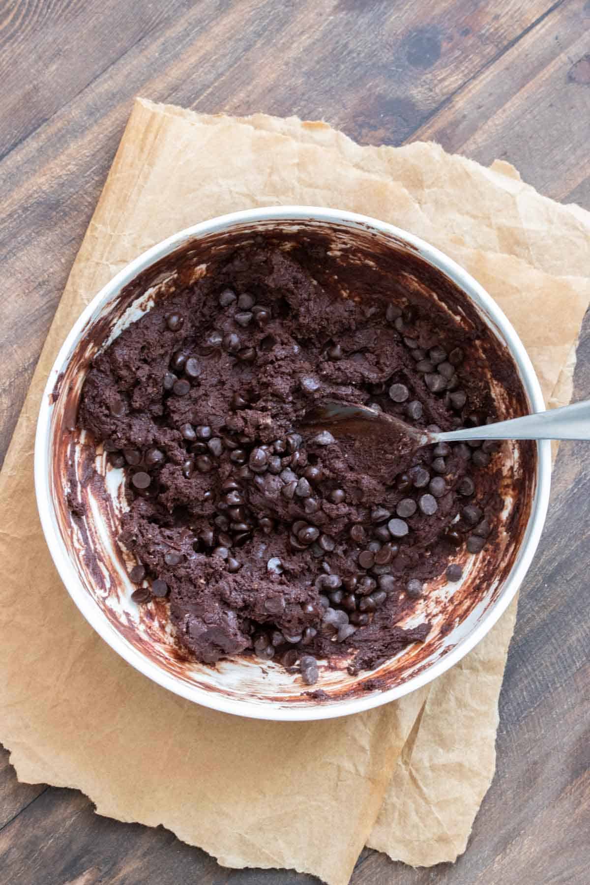 Metal spoon mixing chocolate chips into brownie batter in a while bowl