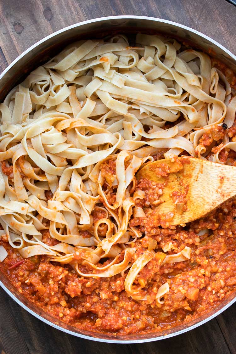 Long flat noodles being mixed into a meaty looking red sauce by a wooden spoon