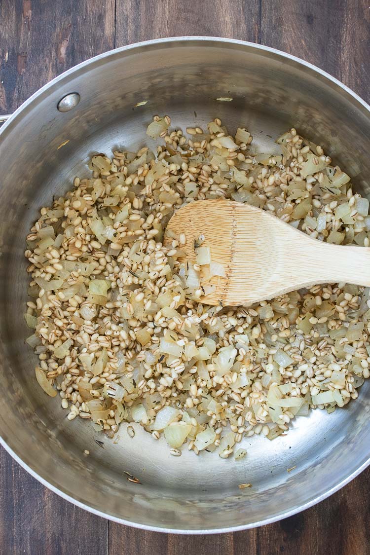 Wooden spoon mixing white onions and barley as they sauté in a metal pot