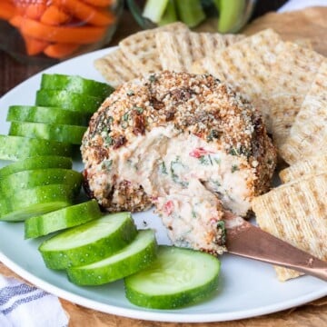 Front view of knife scooping cheese from a cheeseball next to cucumbers and crackers