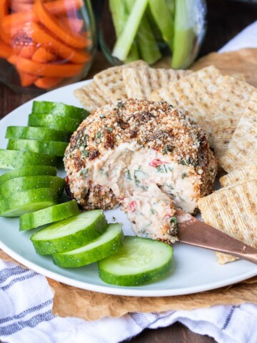 Front view of knife scooping cheese from a cheeseball next to cucumbers and crackers