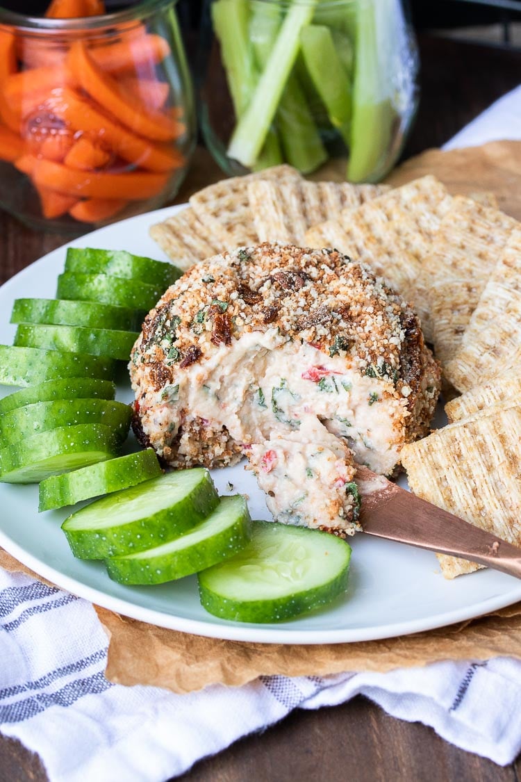 Front view of knife scooping cheese from a cheeseball next to cucumbers and crackers