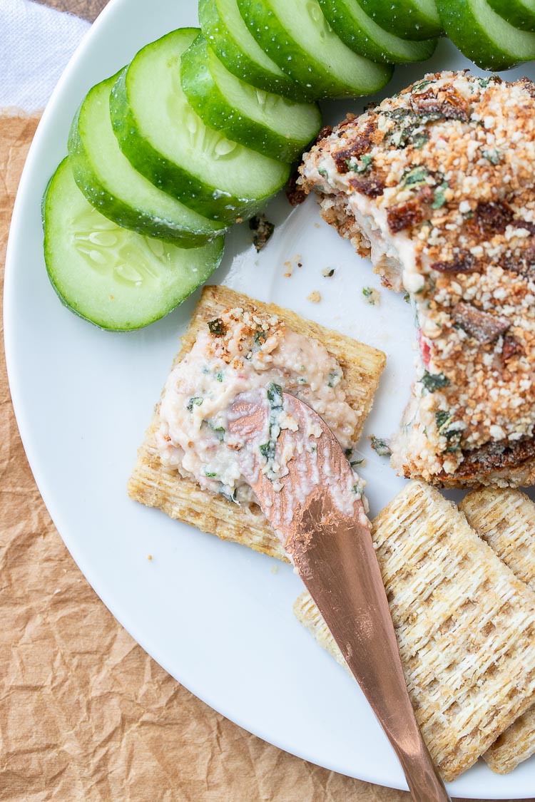 Knife spreading cheese from cheeseball on a cracker next to sliced cucumbers
