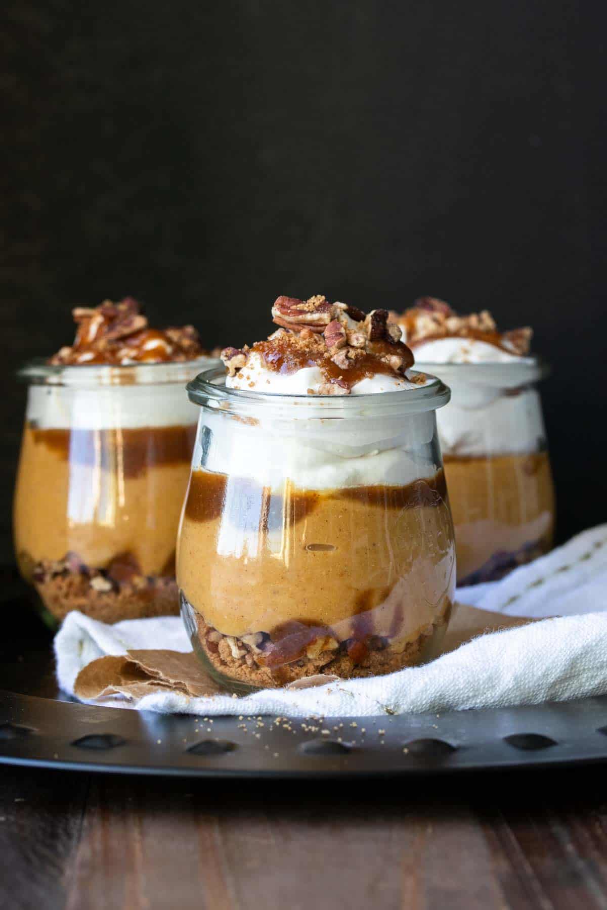 Three glass jars layered with a pumpkin dessert with whipped topping, caramel and pecans.