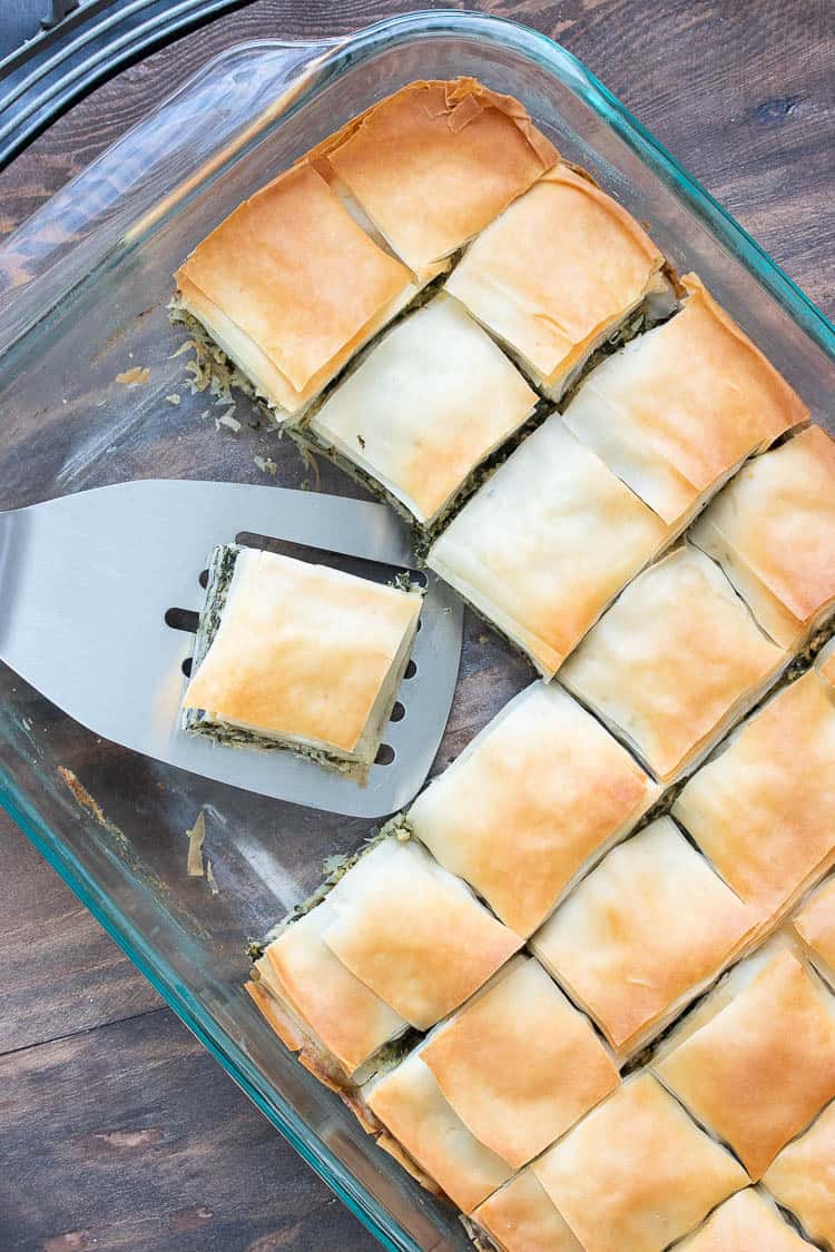 Spatula picking up a piece of baked spanakopita from a glass baking dish