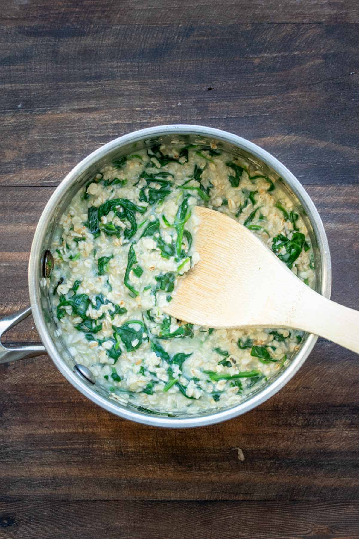 A wooden spoon mixing spinach and oats in a metal pot.