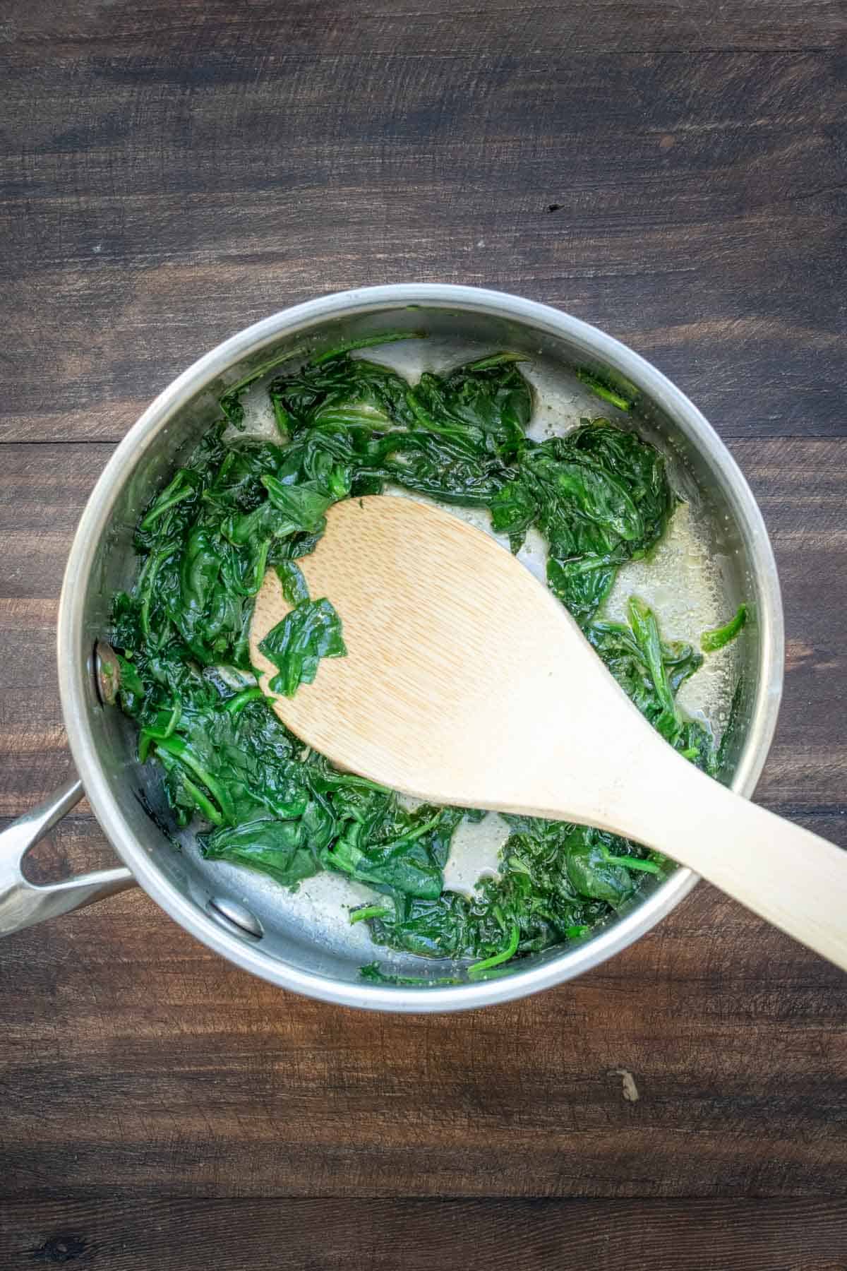 Spinach being sautéed with a wooden spoon in a metal pot.