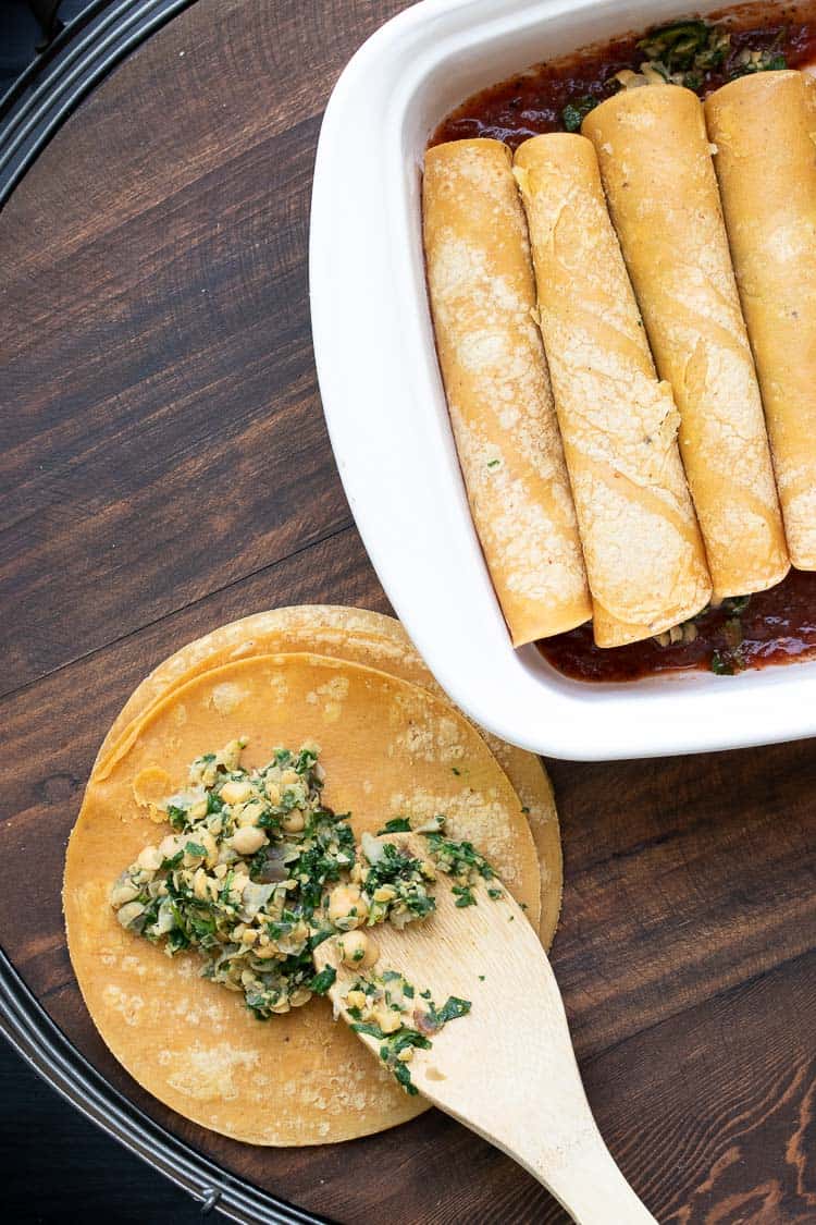 Spoon putting filling on corn tortillas next to a baking dish of enchiladas