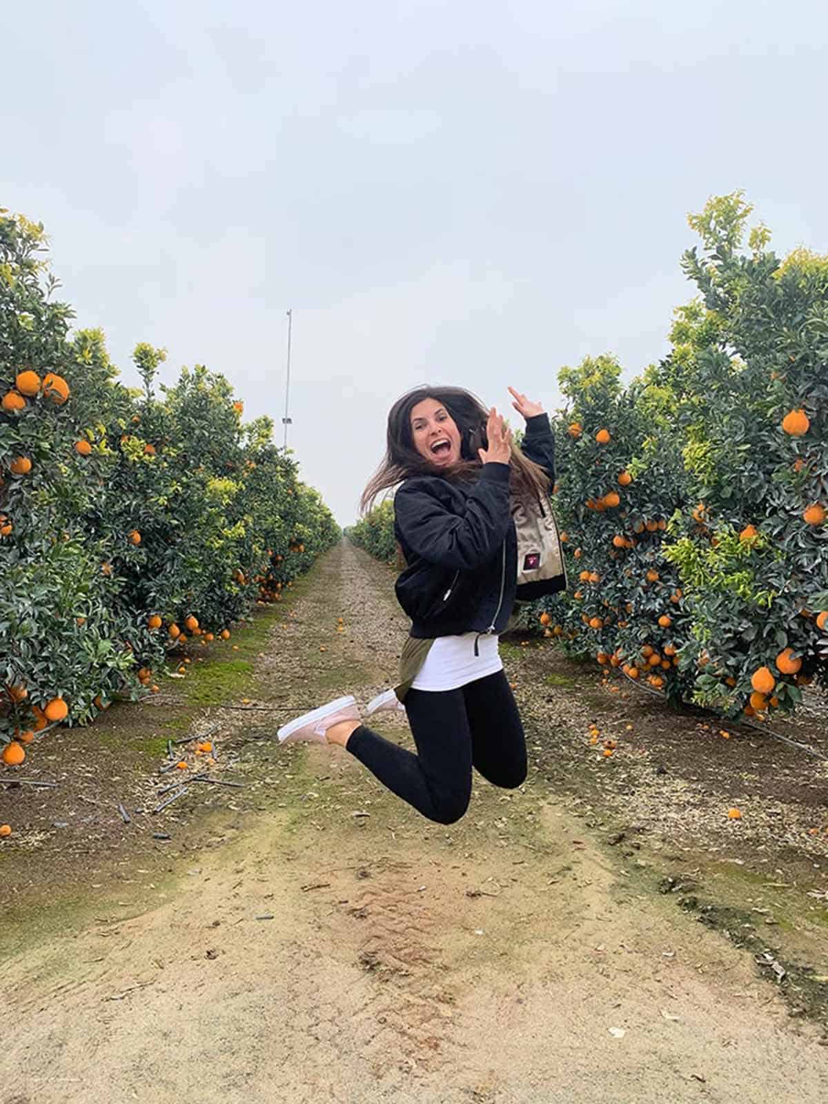 Woman jumping in the air in between two lines of citrus trees