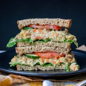 Black plate with a pile of two halves of a mashed chickpea salad sandwich on wheat bread.