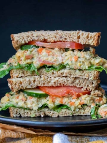 Black plate with a pile of two halves of a mashed chickpea salad sandwich on wheat bread.