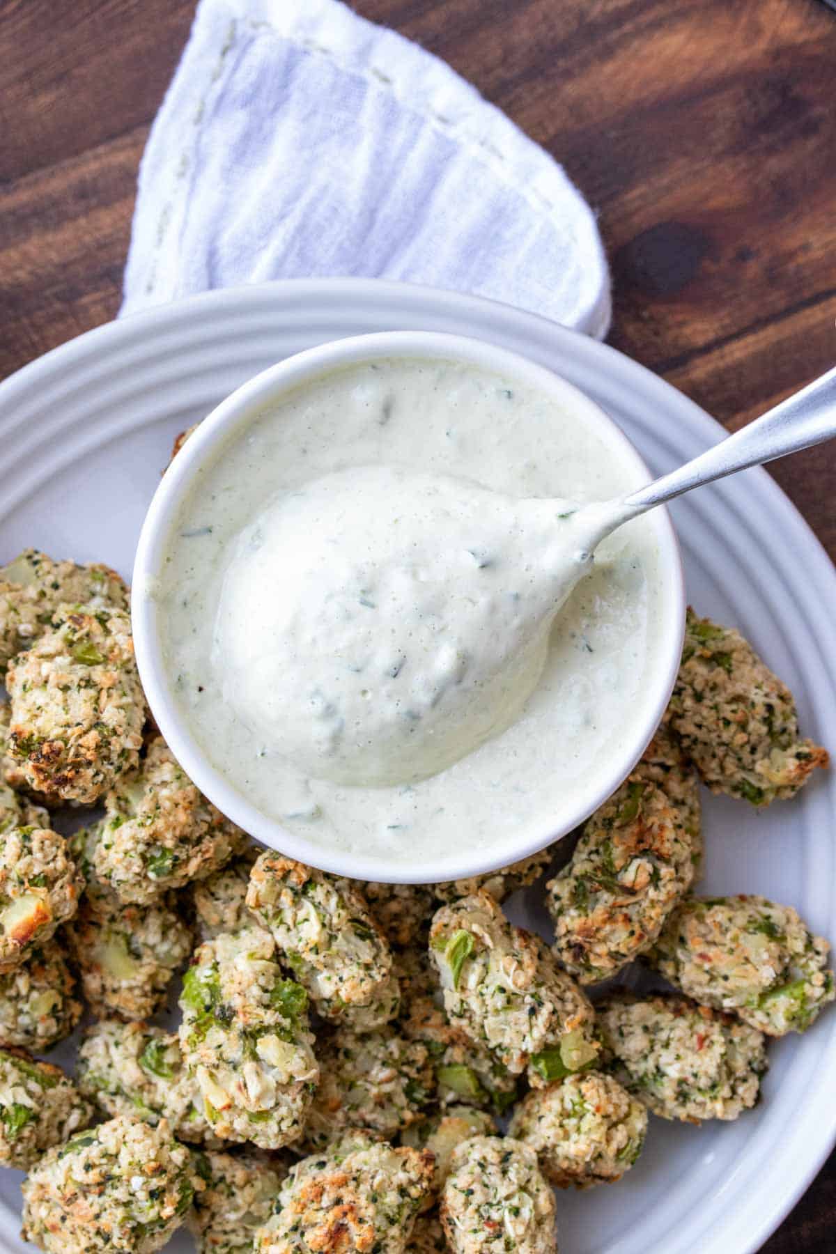 Spoon scooping some creamy dip out of a white bowl next to veggie tots.