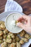 A hand dipping a veggie tot into a white dipping sauce on a plate surrounded by more veggie tots.
