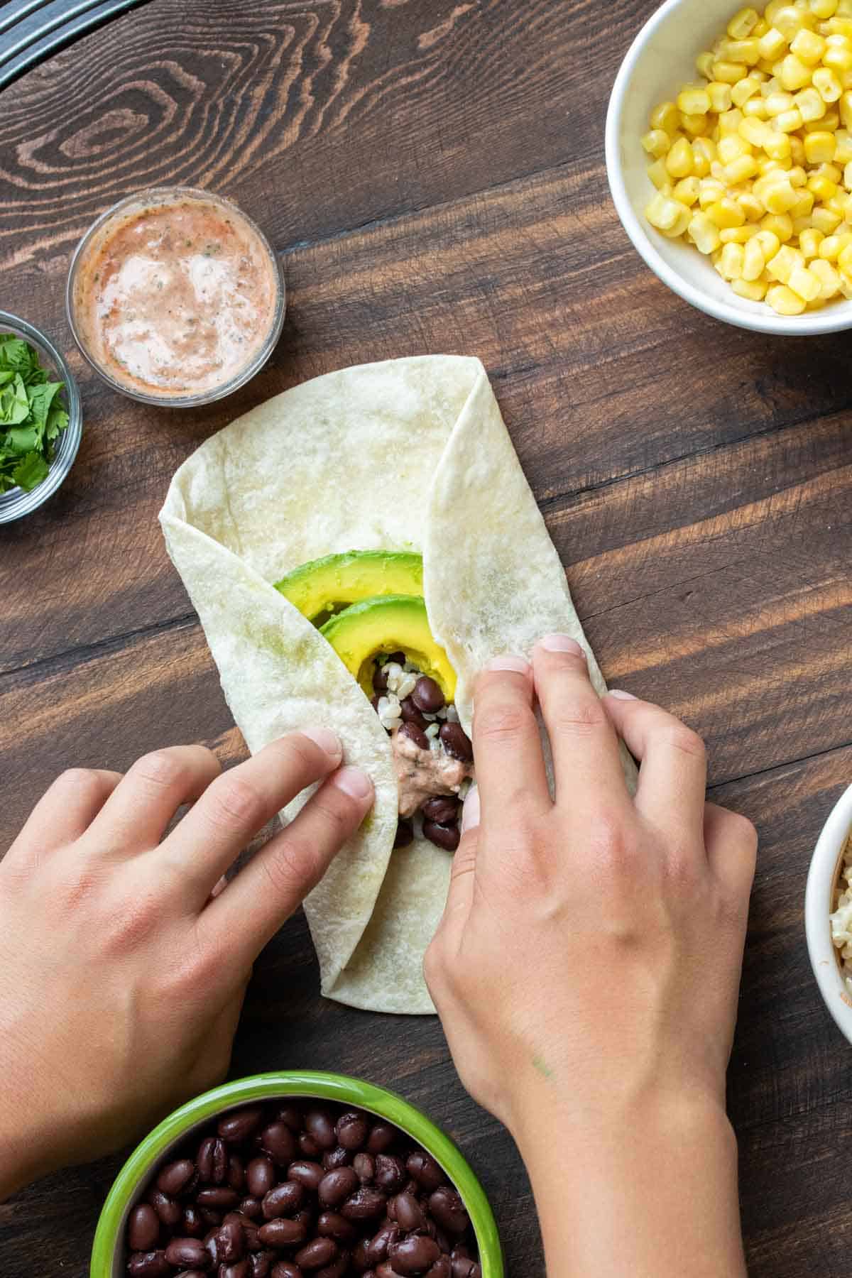 Hands folding the sides of a tortilla with burrito fillings inward