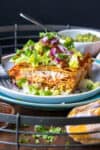 A layered enchilada casserole topped with lettuce and onion on a white plate over a blue plate.