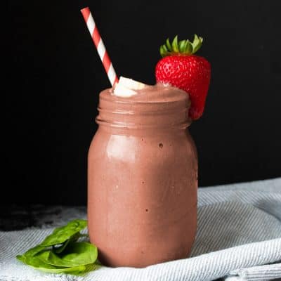 Glass jar with a reddish smoothie surrounded by pieces of strawberry, banana and spinach