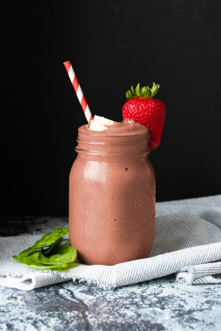 Glass jar with a reddish smoothie surrounded by pieces of strawberry, banana and spinach