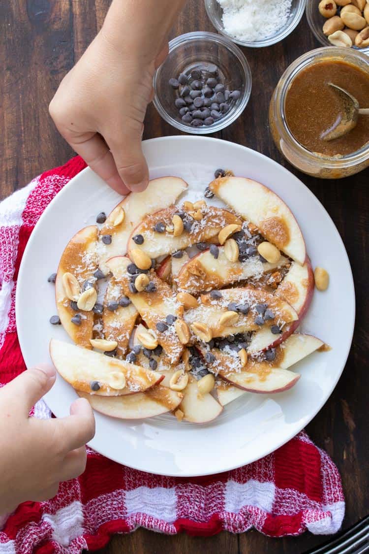 Two hands grabbing apple nachos with caramel and toppings