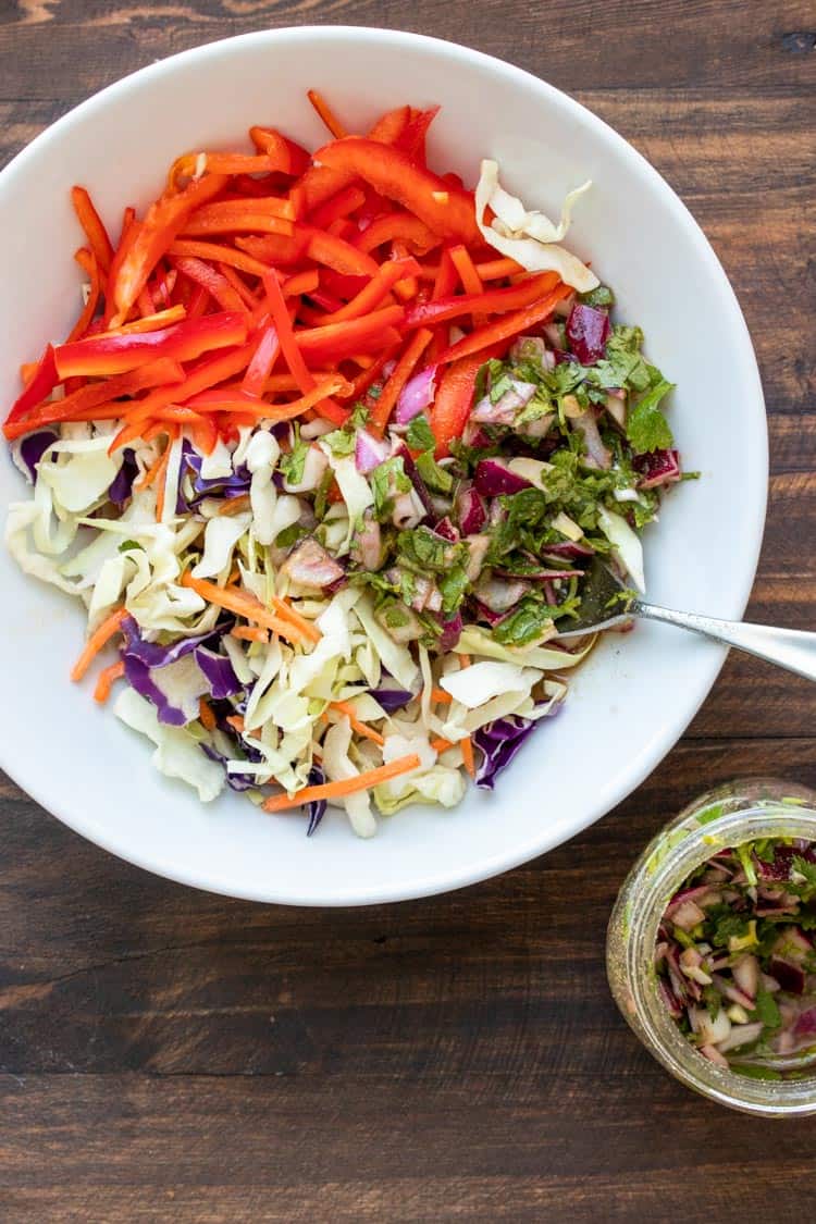 Spoon dipping into white bowl with sliced red pepper, cabbage, onions and cilantro