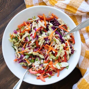Two silver utensils in a white bowl with veggie coleslaw