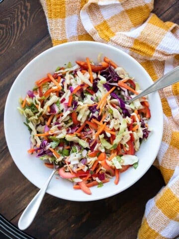 Two silver utensils in a white bowl with veggie coleslaw