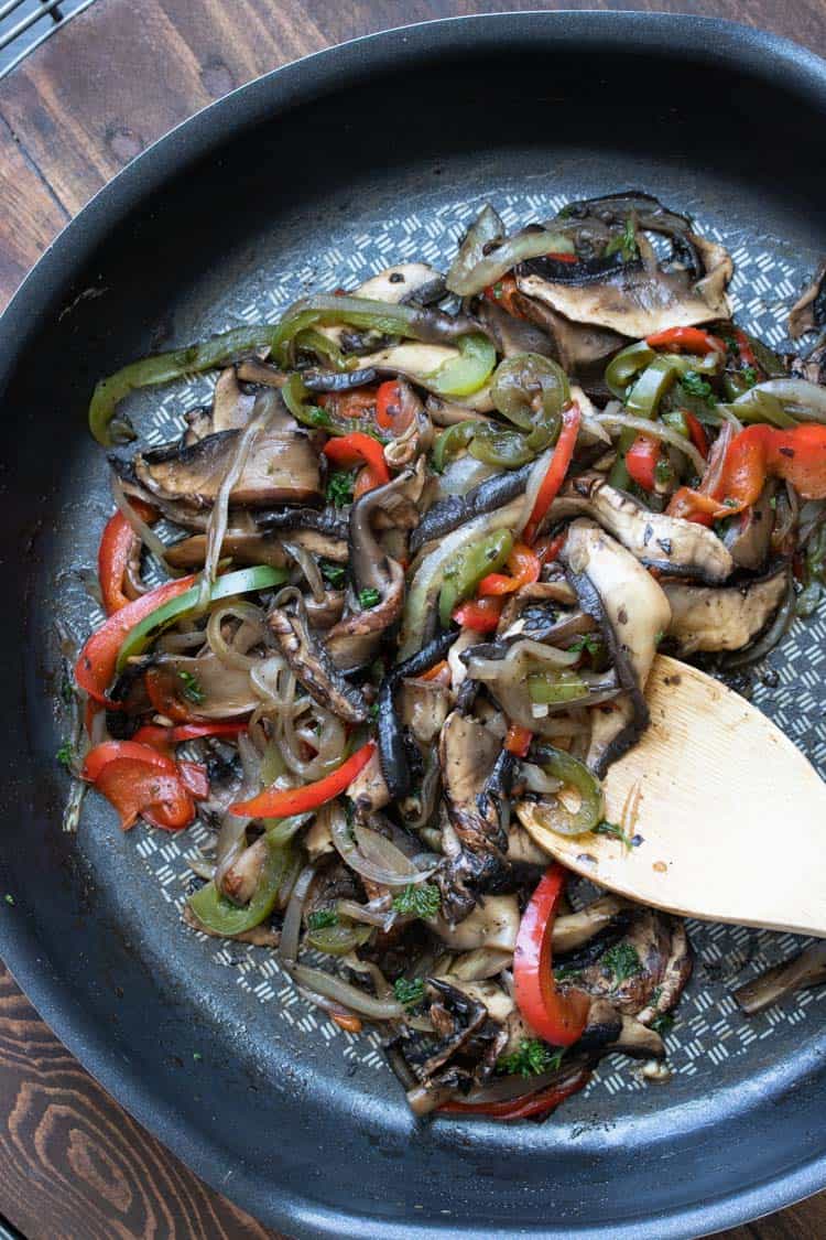 Wooden spoon sautéing mushrooms and peppers in a frying pan